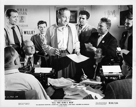 A man wearing a tie around his neck stands in the middle of a room, surrounded by reporters.