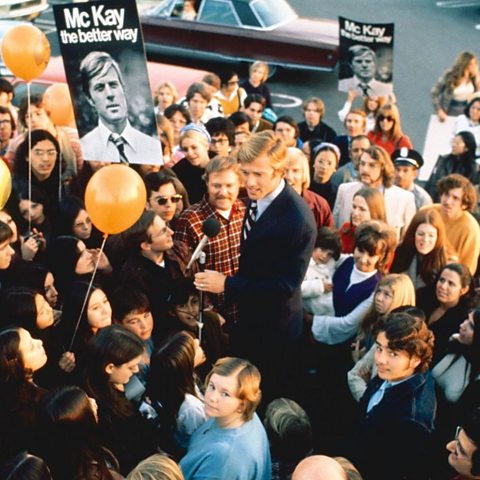 A man with blonde hair, wearing a suit and holding a microphone, makes a speech in the middle of a crowd carrying placards and balloons 