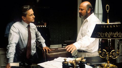 Two men wearing shirts talk to each other behind a desk holding an ornate lamp