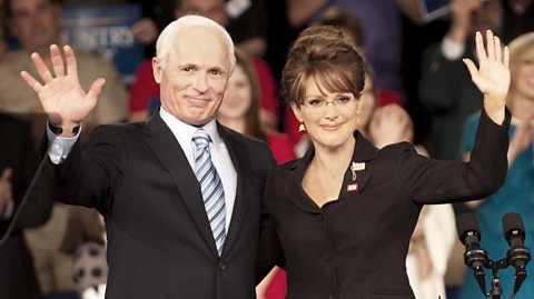 A man and a woman, both suited, wave at a crowd from behind a lecturn, arm in arm