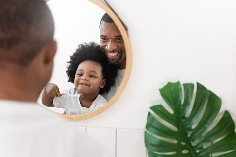 Little boy looking in the mirror with his father holding a white toothbrush 