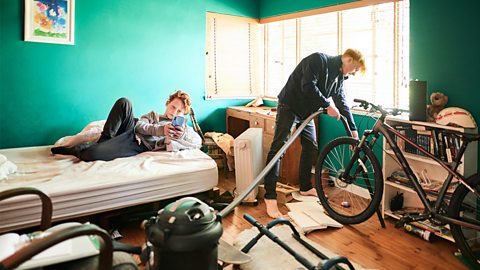 Teenager hoovering his bedroom with his younger brother laying on the bed and looking at his phone