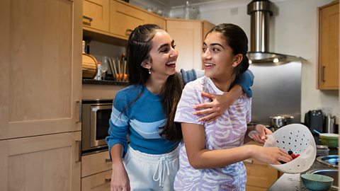 Two sisters cooking together in a kitchen. They are laughing together while the older sister has her arm around the younger. The younger girl is putting berries on a plate.