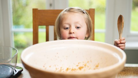 Young girl learning how to prepare a mixture in a large mixing bowl at a kitchen table.