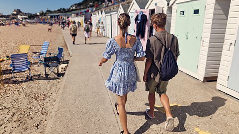 Two tennagers walking next to a row of beach huts, with the beach on the other side