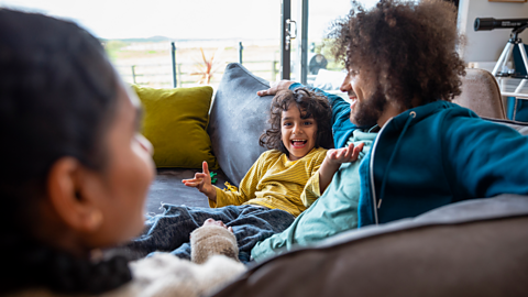 A young boy talks happily slouched on the sofa, his dad cuddling next to him and his mum in the foreground