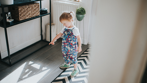 A little girl in flowery dungarees points inquisitively at a shadow on the floor.