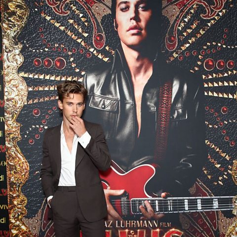 Actor Austin Butler standing in front of a poster of his film Elvis, where he is photographed in the role of musician Elvis Presley, holding a red guitar.