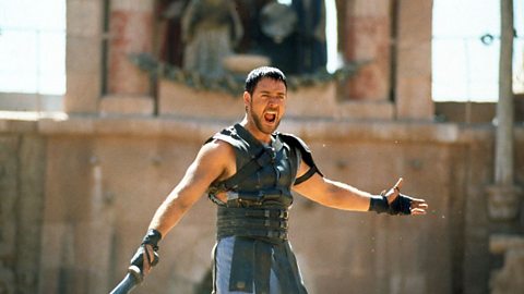 Actor Russel Crowe dressed as a gladiator, handling a sword and with his mouth open in a scream.