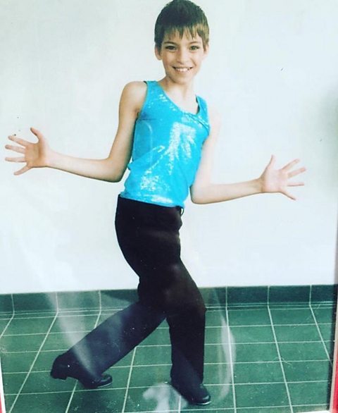 A young boy wearing black trousers and shiny blue leotard does jazz hands