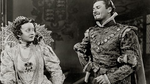 Actors Bette Davis and Errol Flynn dressed as Tudor characters, wearing sumptuous costumes. Hynn is smiling, showing a perfect smile.