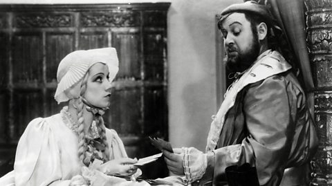 Actors dressed as Henry VIII and his wife Anne Of Cleves, playing cards, dressed in old-fashioned pyjamas.