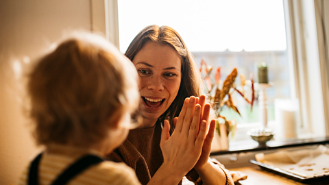 A mother smiles encouragingly at her young toddler while clapping her hands together.