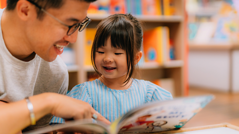 A father wearing glasses smiles and points to something in the picture book while his daughter looks on, holding the book open.