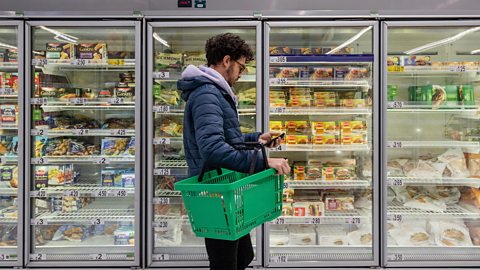 Man shopping in a supermarket. He is standing looking at his phone in front of a row of freezers. 