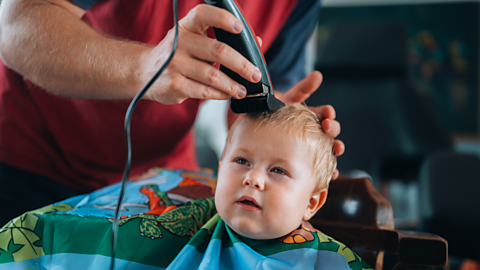 A young boy wearing an apron squints while his dad runs clippers over his head.