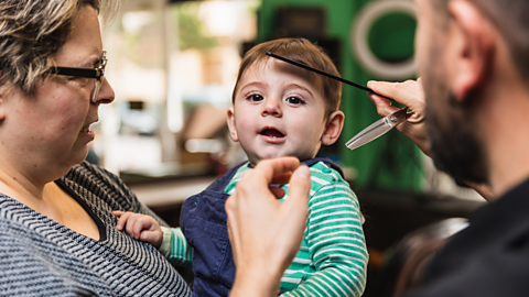 A young boy smiles while being held by his mum, a hairdresser runs a comb through his fringe.