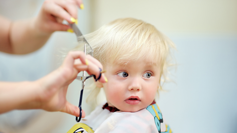 A baby looks nervously off to one side while their hair is trimmed using a comb and scissors.