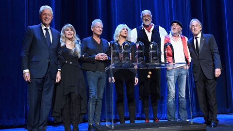 Bill Clinton stands alongside members of Fleetwood Mac on stage, with glass awards sat on a podium