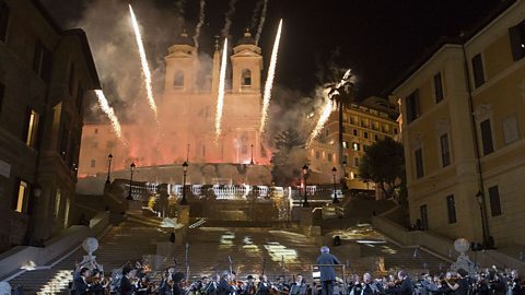 An orchestra playing with Rome's Spanish steps in the background. Yellow fireworks fly up behind them.