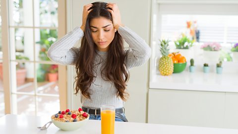 Woman stands with a bowl of cereal and fruit and a glass of orange juice in front of her while she holds her head with a headache