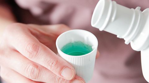 A pair of hands holding a bottle of mouthwash its cap, filled with green liquid.