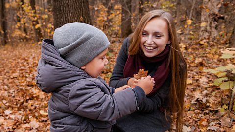 A young boy looks at a fallen oak leaf while his mum looks on encouragingly.