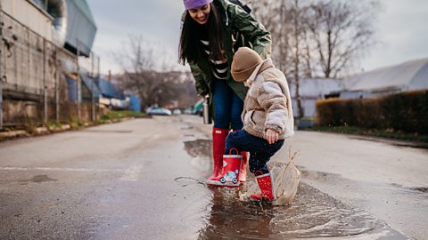 A young child in red wellies splashes in a puddle, their mum looking on.