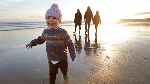 A child in pink fluffy hat runs down the beach, a sunset frames their family in the background.