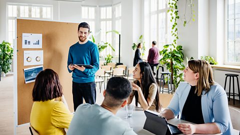 Four people taking part in a marketing focus group. A man in a blue jumper stands in front of the group with graphs pinned to a cork board behind him.