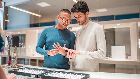 two men who earn large income buying expensive jewellery. One man is wearing a blue top and glasses, the other is wearing a white top. They are shopping for rings.
