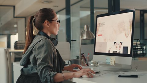 A lady with a pony tail hairstyle and glasses sitting at a computer designing a website.