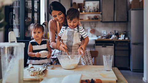 A mother and her two young children have fun in the kitchen, mixing dough and looking through their family recipe book.
