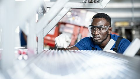A male Quality Control Engineer wearing blue overalls and safety glasses, inspecting steel pipe material for production on shelf rack at factory warehouse
