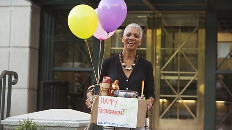 A lady with short grey hair carrying a cardboard box that says Happy Retirement. there are three balloons coming from the box that are purple pink and yellow.