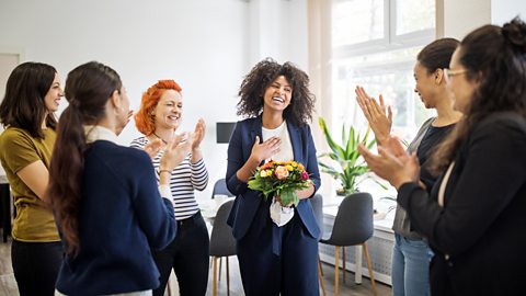 Team of women clapping a colleague who has just got a promotion. She is holding a bunch of flowers.