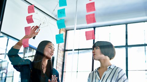 Two ladies working with post it notes on a glass wall to develop a product as their business is growing.