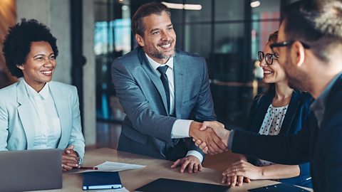 Four business people sit at a table smiling. Two are shaking hands because of a successful business merger