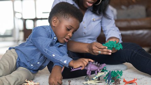 A young boy in a shirt plays with toy dinosaurs, his mum joins in.