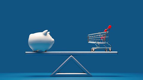 A white piggy bank and a model shopping cart on either side of a balancing scale. The scales are balanced