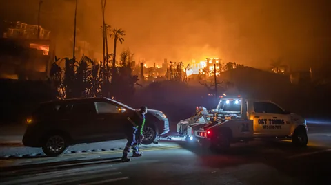 Getty Images An abandoned car is hitched to a tow truck while buildings burn in the background (Credit: Getty Images)