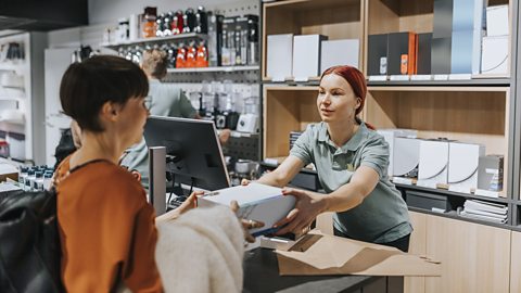 Female customer giving appliance box to sales clerk at checkout counter in electronics store