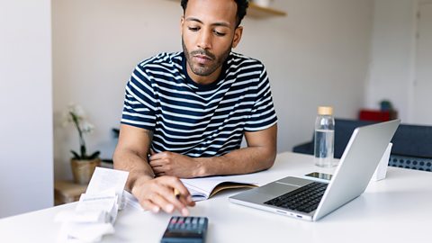 Man sitting at a desk using a calculator and a laptop to calculate his net assets