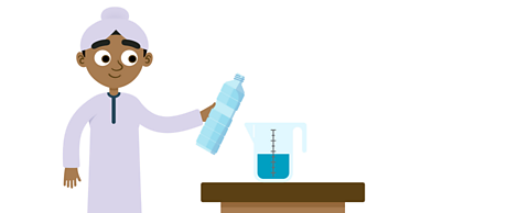 Amir smiling and holding a bottle of water. He is stood next to a table with a jug on it. The jug is half full of water.