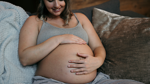 A woman in the third trimester of pregnancy sits on the sofa, holding her bump and smiling