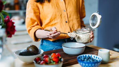 A person puts spoons of porridge oats into a bowl with other bowls filled with fruit (avocado, strawberries, blueberries) near to her