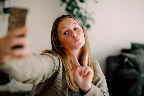 A young girl taking a selfie on her phone. She is pouting and making a peace sign with her fingers. 