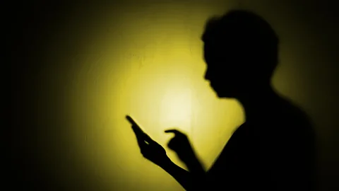 Getty Images A silhouette against a yellow light of a person about to press down on a phone screen (Credit: Getty Images)