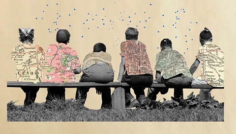 Javier Hirschfeld/ BBC Friends sit on a bench wearing different coloured maps on their shirts indicating they come from different countries (Credit: Javier Hirschfeld/ BBC)