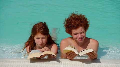 Redheaded girl and boy lying side by side in a swimming pool reading books.